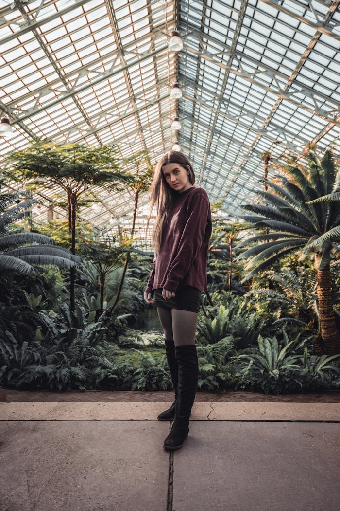 A woman poses stylishly in a vibrant greenhouse surrounded by lush plants.